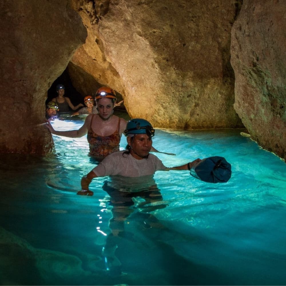 A group of people wearing helmets wade through blue water in a cave.