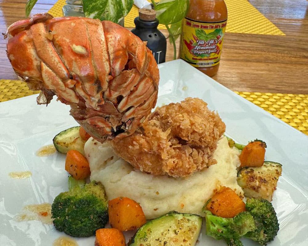 A lobster tail atop a mound of fried chicken, served with seasoned vegetables and mashed potatoes.