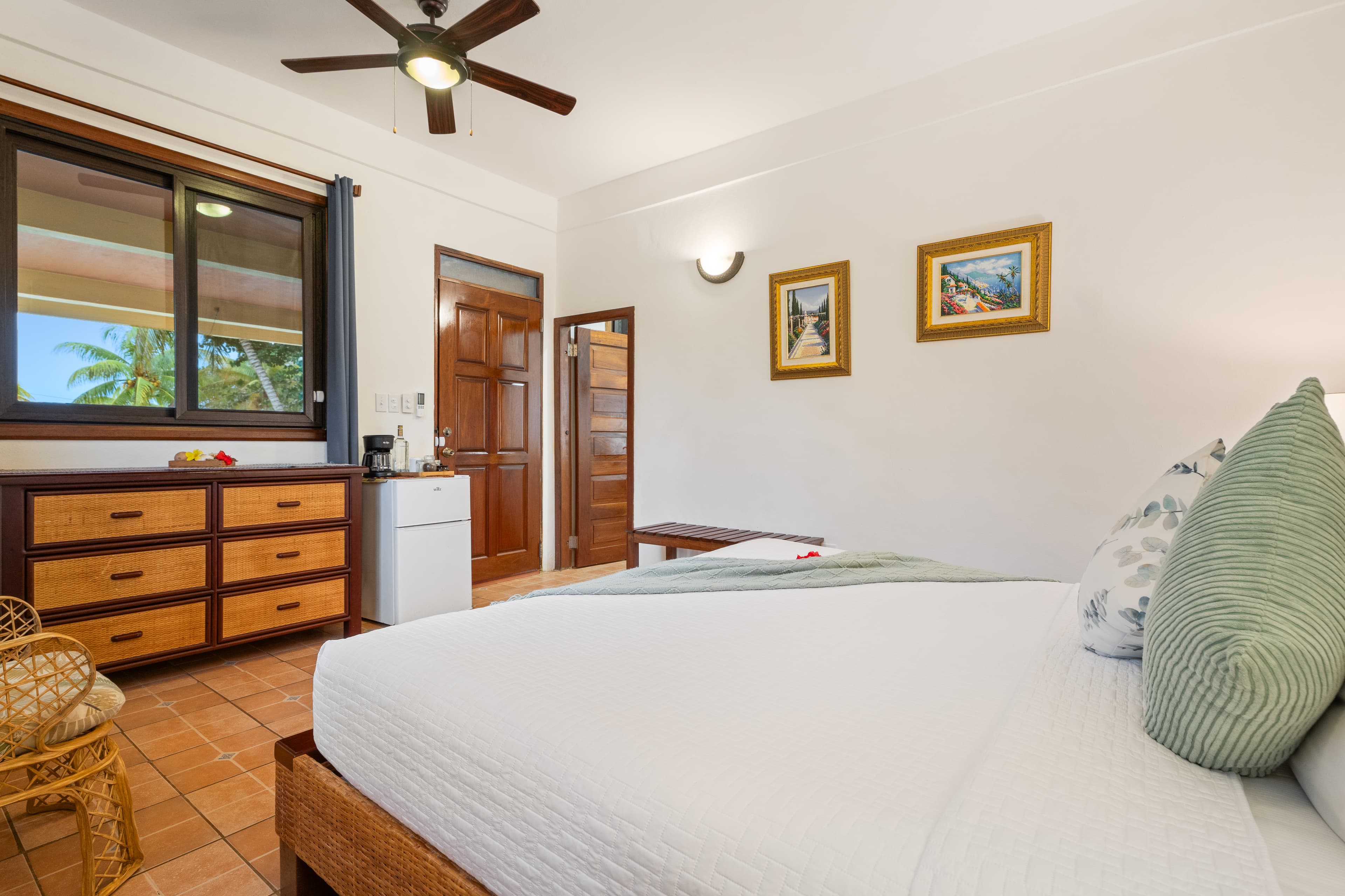 A bright hotel room with terracotta tile floors featuring a king-size bed in the foreground, a wooden dresser beneath a large window, a mini-fridge, and two framed tropical art pieces on the white wall.