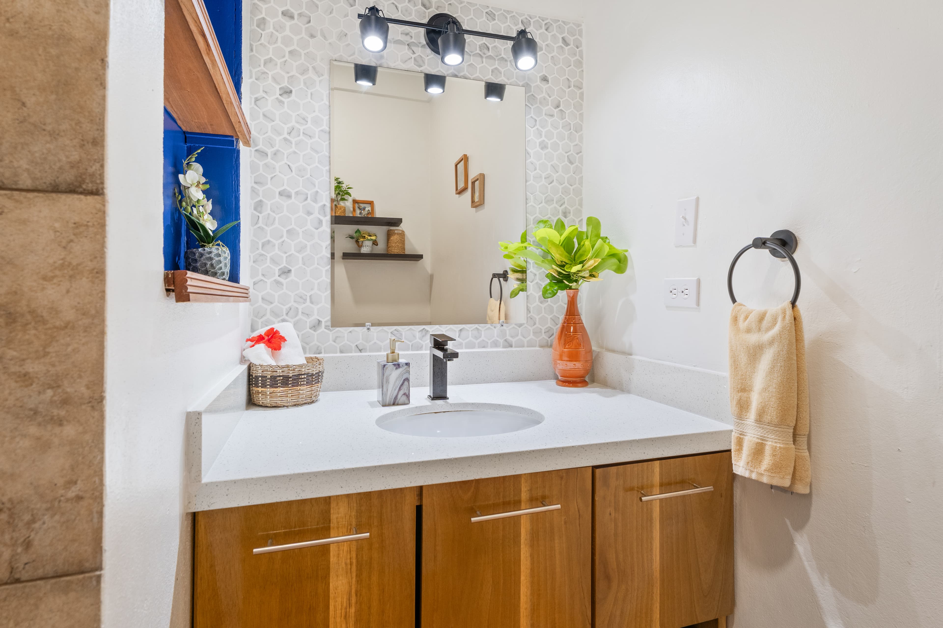 A modern bathroom vanity with a white countertop and wood-grained cabinets, featuring a mirror framed with mosaic tiles and illuminated by a three-bulb vanity light. The space is decorated with a vibrant orange vase holding green foliage, a small woven basket, and a yellow hand towel hanging on a dark circular ring.