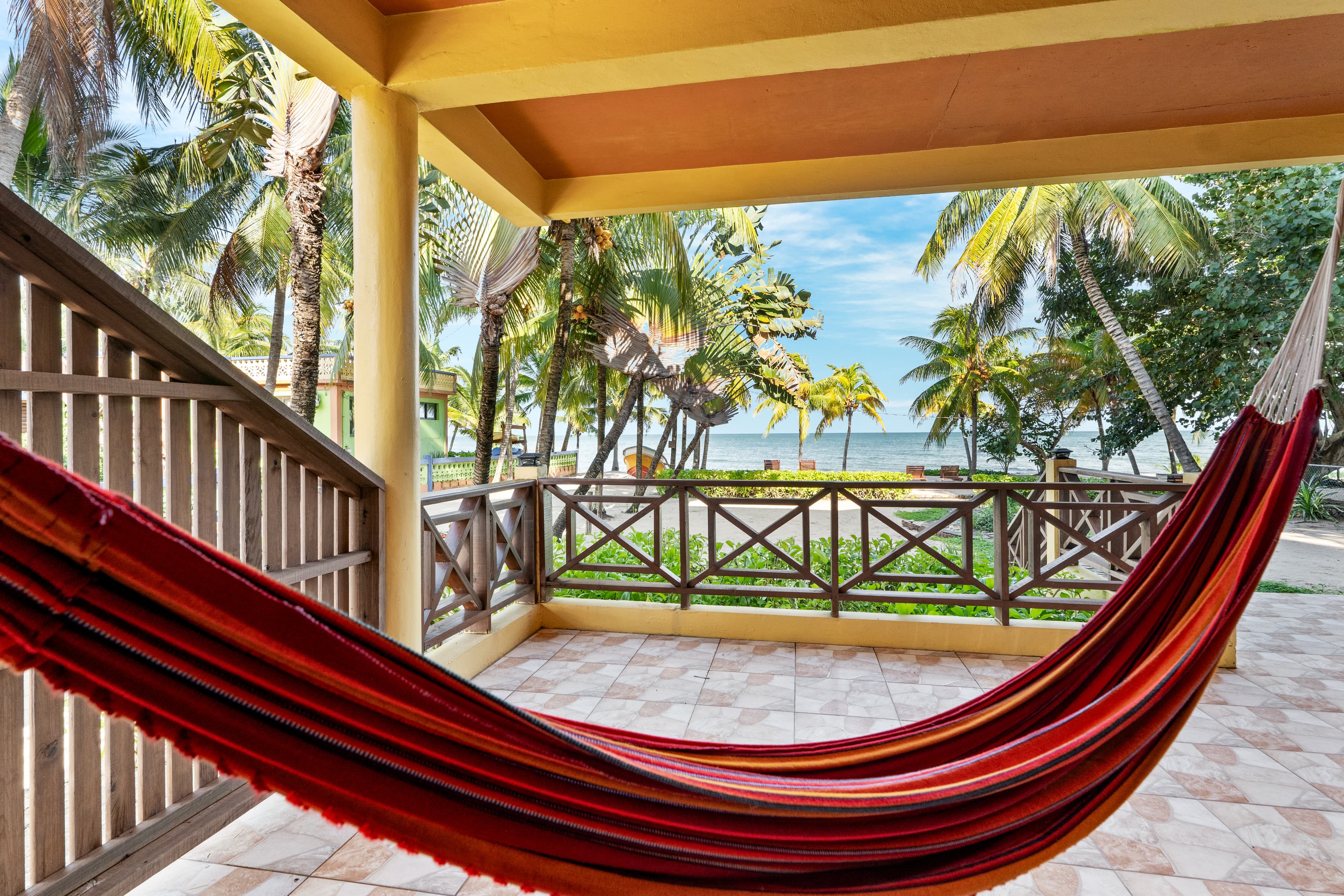 A vibrant, striped red and orange hammock hangs in the foreground of a shaded outdoor patio with tiled flooring. The patio is enclosed by a dark wood railing, offering a clear view of a lush tropical landscape with numerous palm trees and the calm blue sea in the distance.