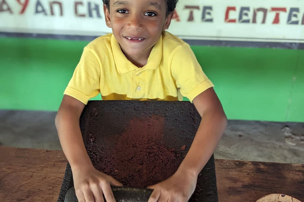 A smiling child in a yellow shirt grinds cocoa beans on a stone slab.
