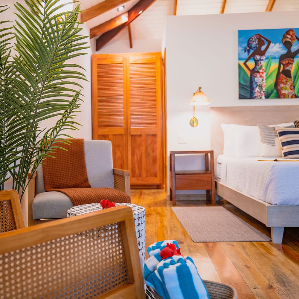 A cozy interior of a room featuring a bed, armchair, and vibrant artwork, with wooden accents and tropical decor.